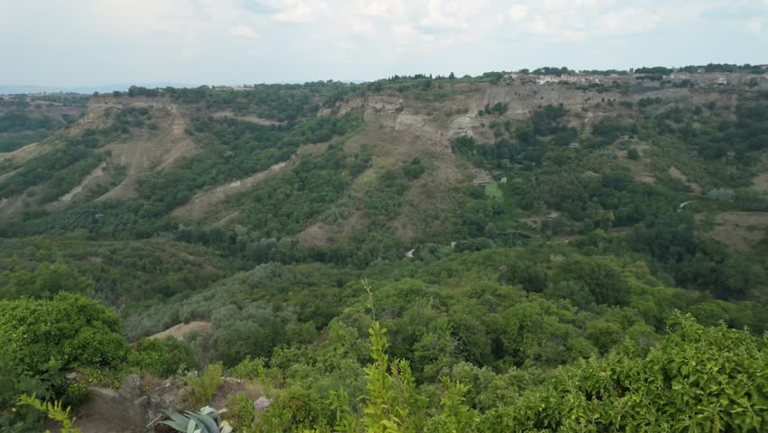 Expansive view of the unique Calanchi badlands, featuring deeply eroded hillsides and vibrant green vegetation, seen from a high vantage point with a foreground garden.