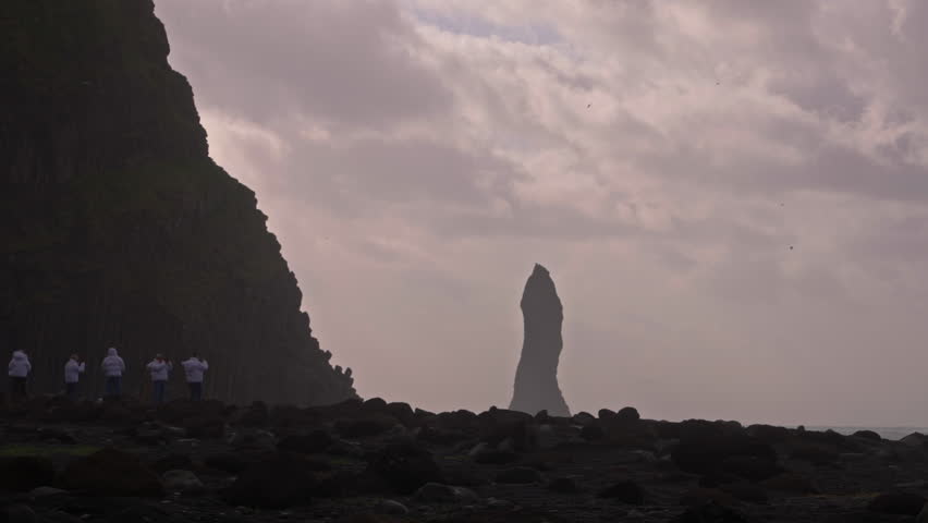 Tourists in white coats walk near basalt columns at Reynisfjara Beach