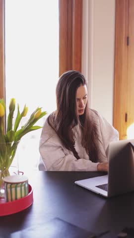 Thoughtful young woman sitting at home office desk with laptop, pensive girl looking away and then smiling at camera, authentic freelancer lifestyle concept, vertical video