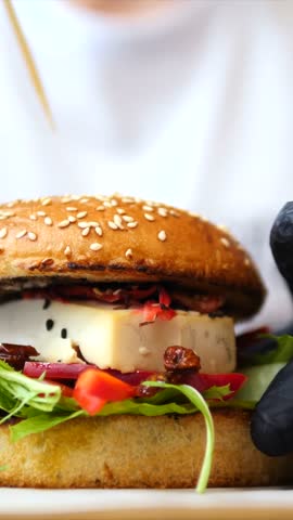 Professional chef in black gloves inserting a wooden skewer into a delicious gourmet burger with cheese, fresh lettuce, and vegetables, preparing the sandwich for serving in a restaurant kitchen
