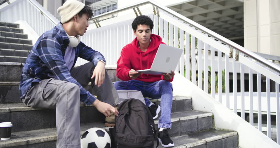 Diverse male friends placing laptop on lap, pulling folder from backpack, studying on stairs. Collaboration, youth, urban, casual, technology, education, teamwork