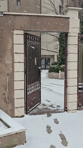 Winter scene with open metal gate and falling snow in a quiet residential yard. Fresh footprints visible on snowy ground, cold seasonal atmosphere and calm outdoor environment