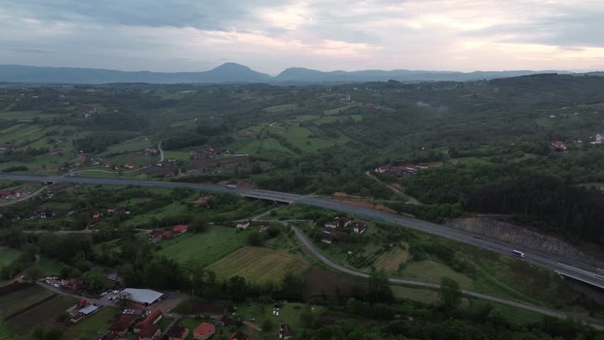 Drone view of mountains with forested hills and highway at sunset with scattered houses. Cars and trucks are driving along an asphalt road among the green spring mountains. Horizontal 4k footage.