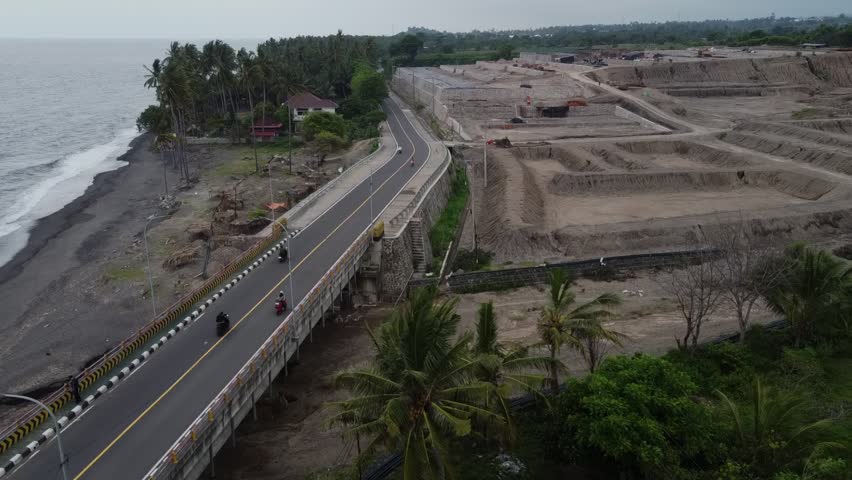 Drone aerial view of a pedestrian bridge near the sea, with coastal water on one side and construction soil piles on the other. Urban development and infrastructure concept.