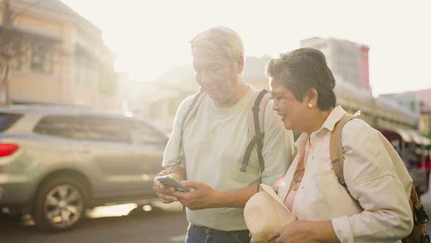 Asian senior couple search direction on phone while exploring the city. Elderly man and woman tourist walk along street, enjoy leisure time urban travel and joyful experience during holiday vacation.