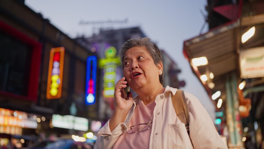 Asian senior woman talking on phone call while exploring city outdoors. Elderly older female tourist walking on street, enjoy leisure time urban travel and joyful experience during holiday vacation.