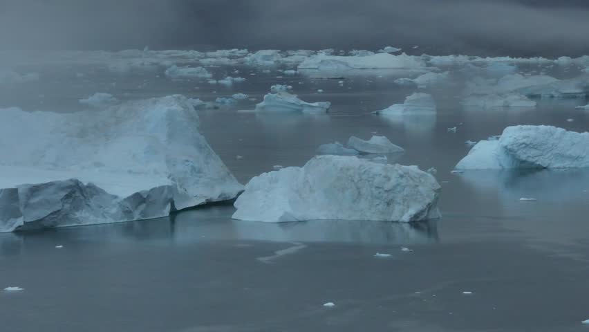 Slow aerial panning across a serene graveyard of crystalline icebergs floating in the misty Arctic waters of Greenland, 4K drone footage.