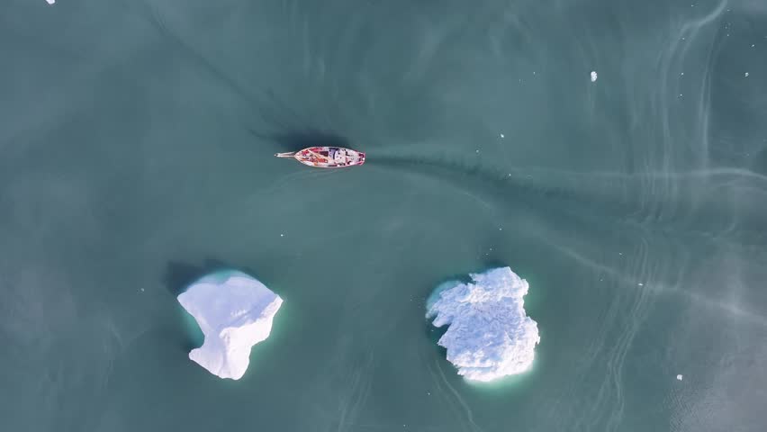 Top-down push-in shot of a red wooden schooner navigating through dark arctic waters past large white icebergs in Greenland, serene atmosphere.