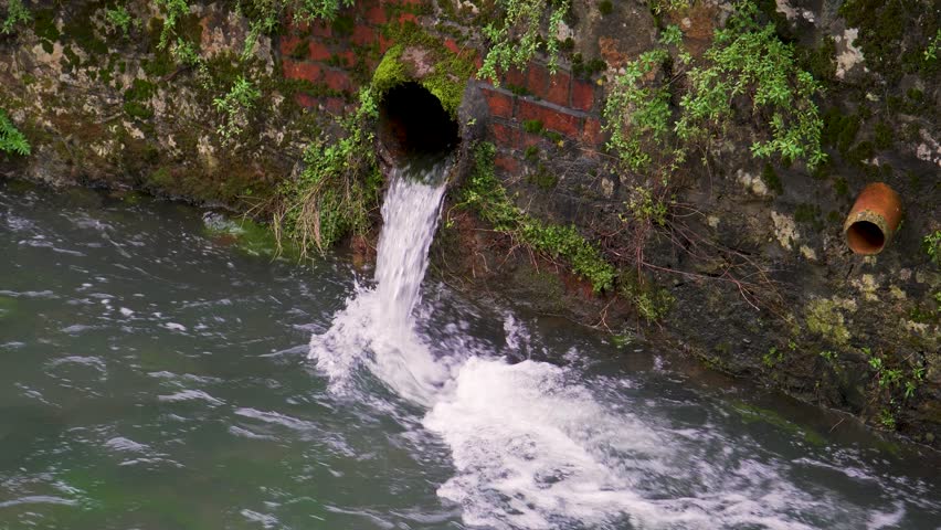 Close up view of one tube from the gutter system coming off one of the red brick wall full of moss and other plants, discharging water into the river Cheddar Yeo. River flowing down hill.