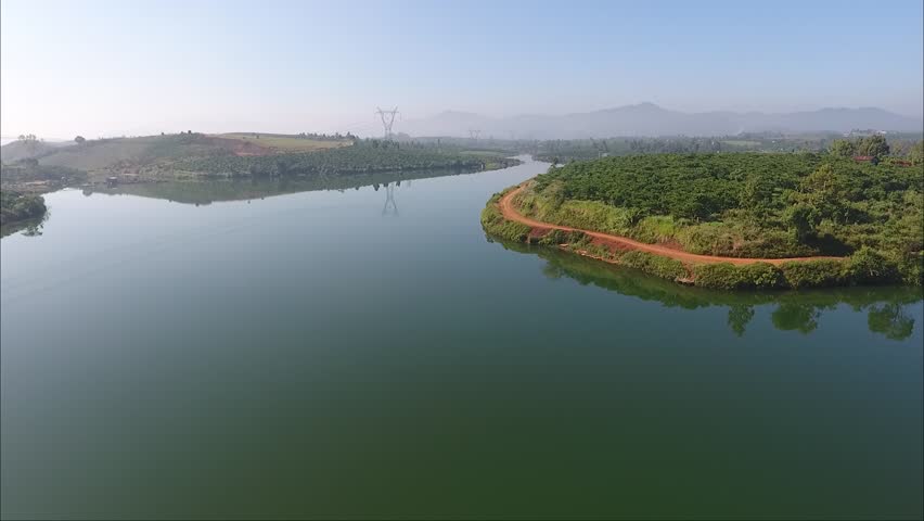 Aerial drone view of rural reservoir with plantation fields and distant mountain range. Agricultural countryside landscape with water surface and green farmland.