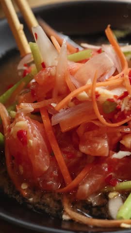 Close-up vertical shot of chopsticks stirring raw salmon, carrots, and chili in a bowl.