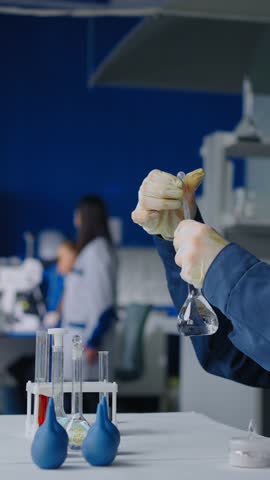 Scientist wearing protective gloves conducts precise liquid experiment in modern laboratory, carefully handling glass flasks and test tubes on a white workbench