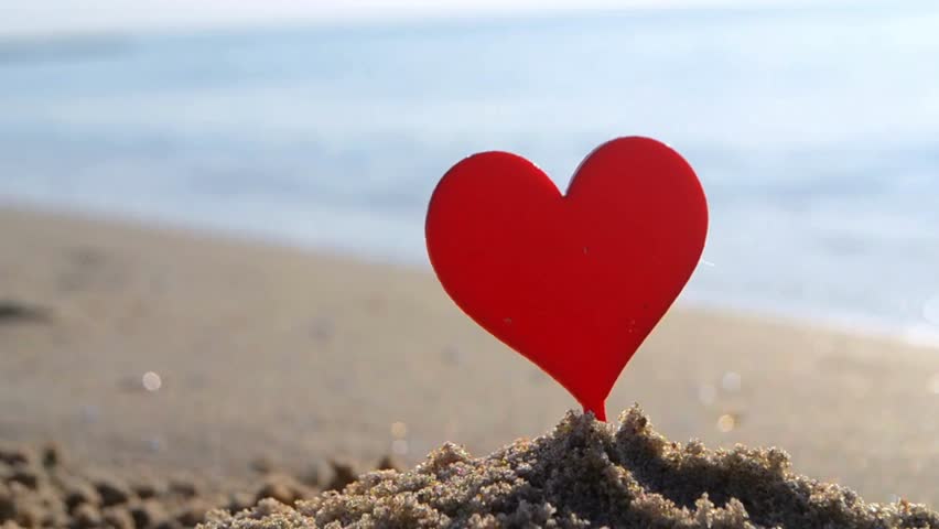 Plastic stick in shape of red heart in sand on sandy beach of sea shore on background of sea waves on sunny summer day close-up. Concept Love relationship romance amorousness amour St Valentines Day