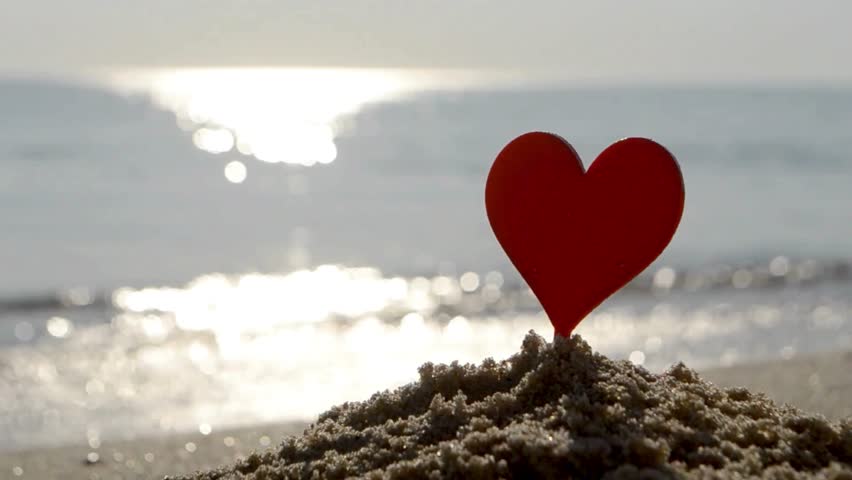 Plastic stick in shape of red heart in sand on sandy beach of sea shore on background of sea waves on sunny summer day close-up. Concept Love relationship romance amorousness amour St Valentines Day