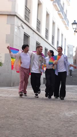 Diverse group of four young friends walking together down a cobblestone street. They are happily celebrating while waving rainbow flags