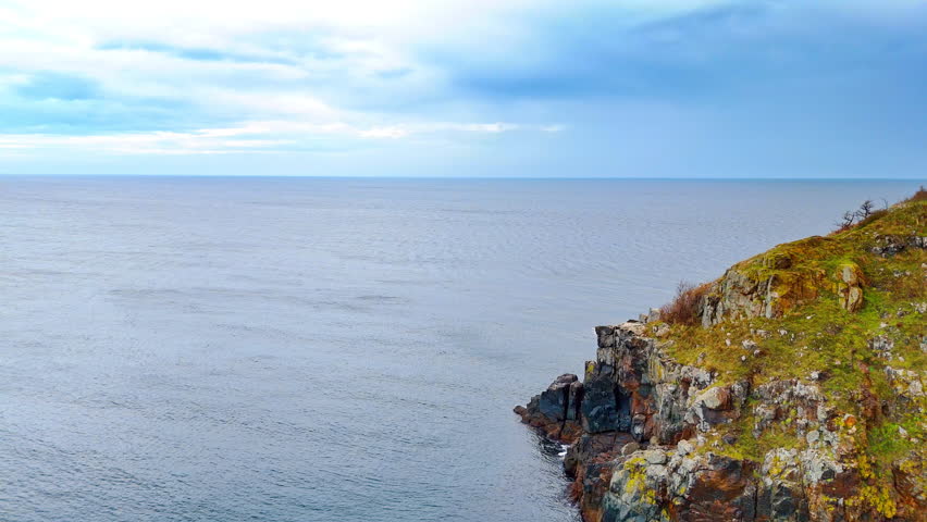 High angle view of rocky cliff edge and vast ocean. Side view of a steep rocky cliff covered with grass overlooking the calm open sea under a cloudy sky.
