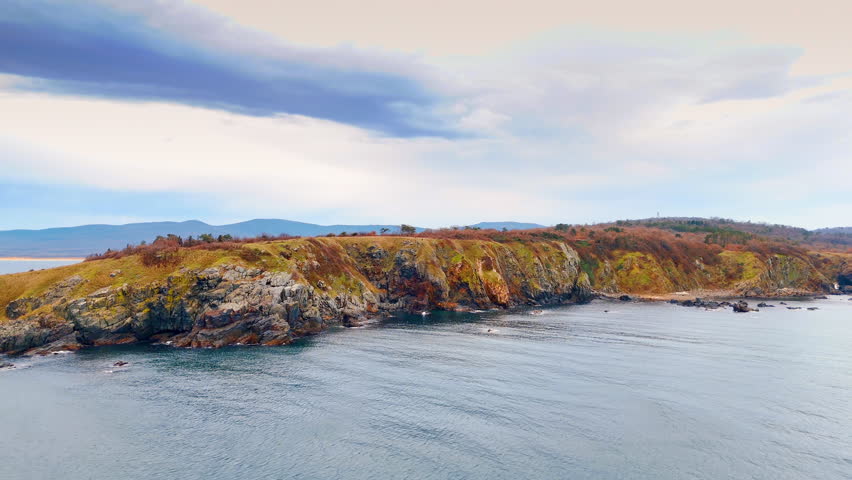 Wide view of rocky coastline and calm sea. Landscape of steep coastal cliffs with autumn bushes meeting the sea.