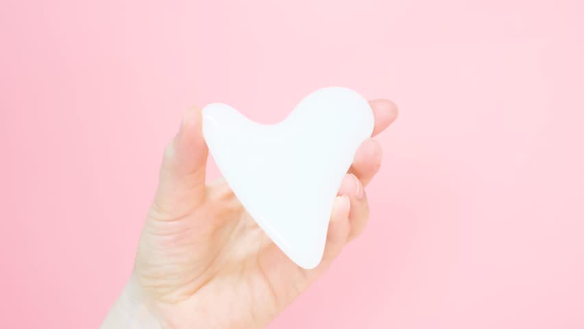 A hand holding gua sha face massager on pink background.
