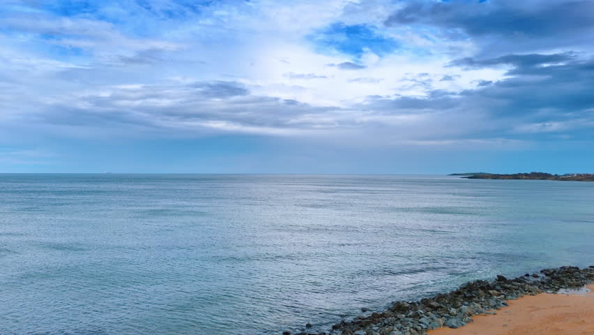 Wide seascape with rocky shore and overcast sky. Calm sea water stretching to the horizon with a stone breakwater in the foreground.