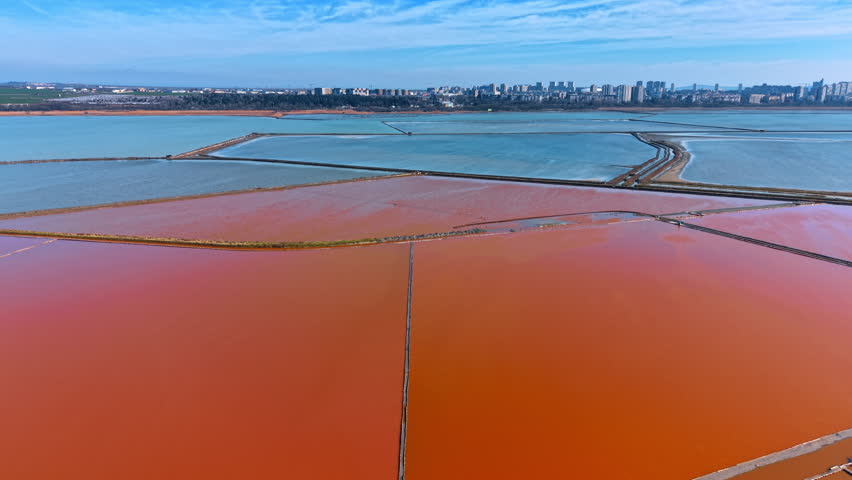 Panorama of pink and blue salt evaporation ponds near city. Wide aerial shot of colorful salt pans with city skyline under clear blue sky in the background.