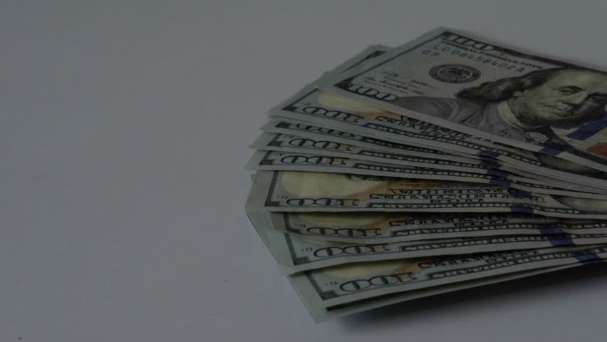 Fanned hundred dollar bills under shadow on white desk, closeup of crisp banknotes with Benjamin Franklin portrait and blue security strip, subtle creeping shadow evokes audit