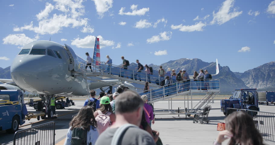 Daytime summer footage of passengers boarding a commercial airplane on the tarmac at Jackson Hole Airport in Wyoming. Boarding takes place outdoors via a ramp, with the Grand Teton Mountains visible in the background. No recognizable faces are visible. The scene highlights air travel, airport ground operations, and one of the most scenic mountain airports in the United States. Ideal for travel, tourism, aviation, and transportation stock footage.