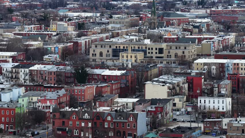 Aerial view of a historic Baltimore neighborhood with colorful rowhouses, brick facades, church steeple, and dense urban blocks under soft winter light. Wide shot. Maryland, USA in winter.