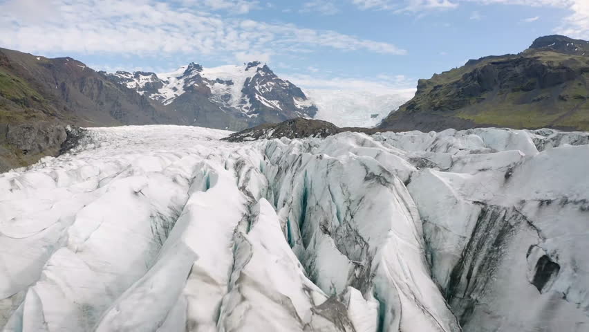This clip showcases a stunning aerial view of a glacier with intricate ice formations and rugged mountains surrounding it. The clear blue sky adds to the serene beauty of the natural landscape.