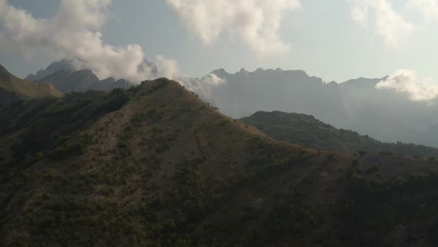 A serene view of a mountainous landscape featuring rolling hills and distant peaks partially shrouded in clouds. The lush greenery contrasts with the rocky terrain, creating a tranquil natural scene.