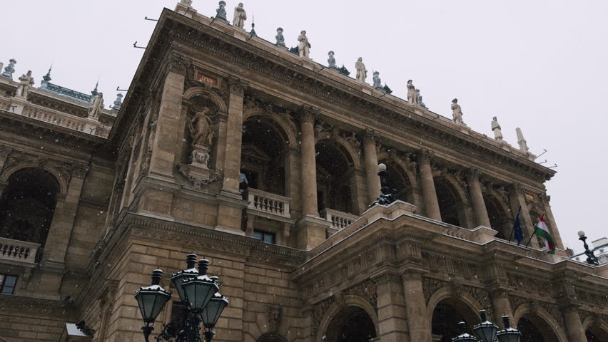 Wide view of the Hungarian State Opera House in Budapest during active snowfall. Historic European architecture, stone columns, statues and winter city atmosphere.