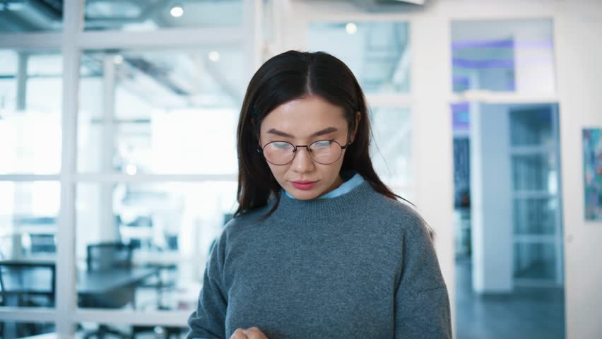 Female employee pausing work on tablet and looking directly at camera. Woman tapping on tablet screen with fingers. Confident expression in glass office corridor. Showing positivity and joy.