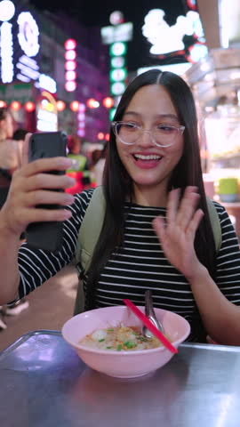 Happy beautiful asian female tourist takes a selfie with her bowl of Thai noodle soup using a smartphone at a vibrant neon-lit street food night market in Bangkok city during her vacation. Vertical Shot
