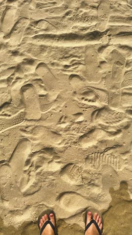 Person standing in flip flops on a sandy beach, showing various footprints, conveying themes of walking, exploration, leisure, and summer relaxation in a warm, outdoor setting