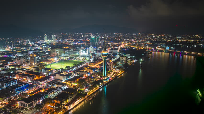 Aerial night view of Kuantan riverside city illuminated with vibrant architectural lighting and flowing traffic. Skyscrapers, urban streets, and a prominent observation-style tower create a dynamic skyline, while reflections shimmer across the calm water. Captured during a time-lapse sequence, the scene showcases motion, energy, and contemporary city living, making it ideal for themes of business, travel, infrastructure, technology, and urban development. Clean, location-neutral composition suitable for global commercial use.