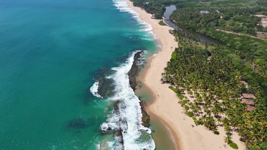 Breathtaking aerial drone footage of the vast coastline in Tangalle, Sri Lanka. The video captures a long stretch of golden sandy beach separating the turquoise Indian Ocean waves from a calm river lagoon. The landscape is filled with dense green coconut palm forests and mangroves