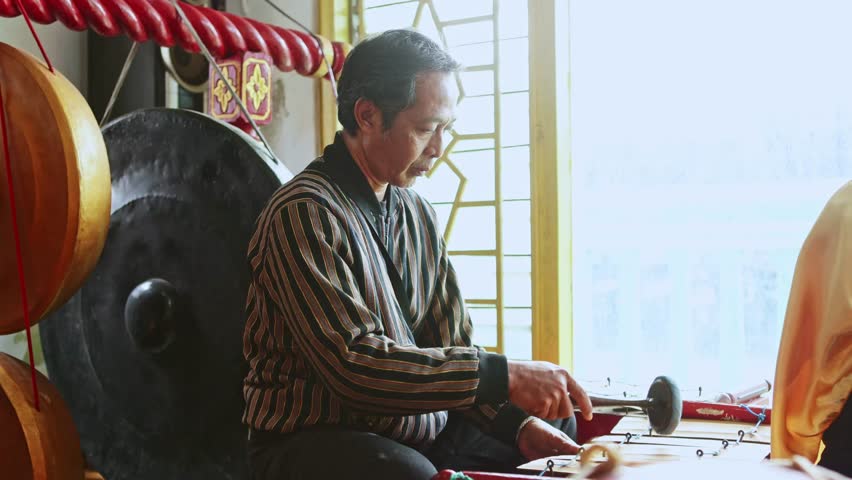 Professional Javanese musician sitting and playing a traditional gamelan ensemble percussion instrument with a mallet during a cultural performance in a music studio with a bright window behind him