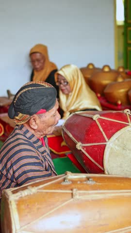 Elderly Javanese musician in traditional batik clothing playing a drum in a gamelan ensemble, surrounded by percussionists during a cultural performance