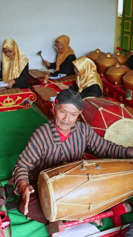 Elderly Indonesian man in traditional clothing skillfully playing a traditional hand drum, as part of a classic Javanese Gamelan musical ensemble performing at a cultural event