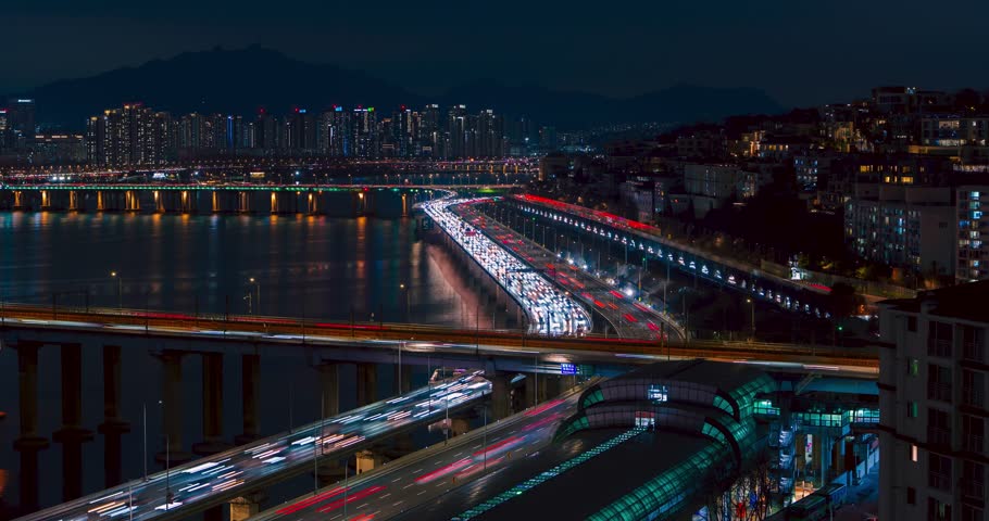 Timelapse displays continuous night traffic light trails streaming across Gangbyeonbuk-ro expressway and Donho Bridge over Han River surrounded by Seoul skyline high-rises and reflections