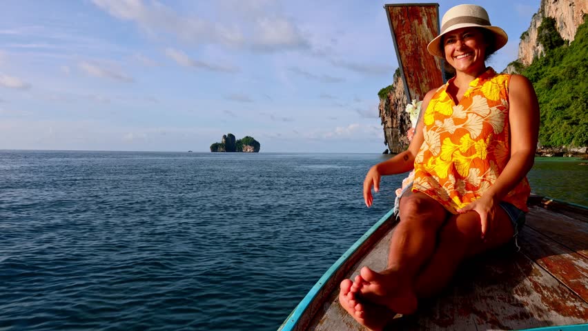 Cheerful female tourist wearing a sun hat and sitting on the bow of a wooden long tail boat, enjoying the stunning sea view of the Phi Phi Islands in Thailand during a sunny day