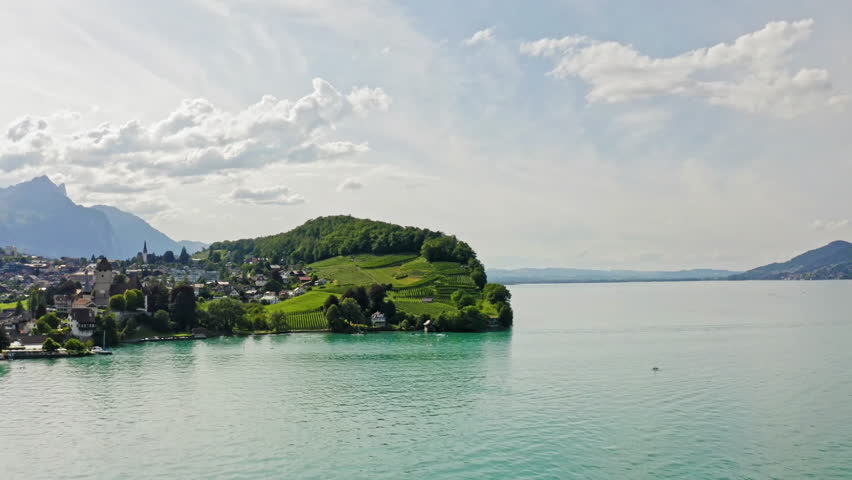 Aerial view of a serene lake surrounded by rolling green hills and a small village, with mountains in the background under a partly cloudy sky.