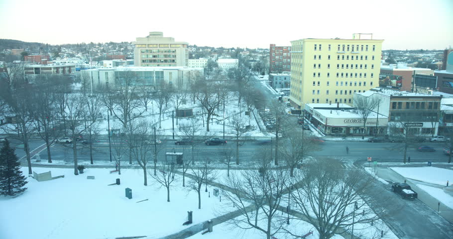 A day-to-night time lapse of downtown Manchester, New Hampshire during the winter. The shot overlooks Elm Street with Veterans Park in the background. As the sun sets, the sky transitions from twilight to dark, and the city lights and street lamps illuminate the snowy landscape. Traffic flows through the main street of the Queen City as the evening commute begins.