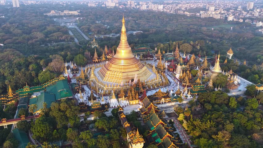 Aerial drone view at Shwedagon Pagoda, a golden stupa, surrounded by lush greenery and urban development in Yangon (Rangun), Myanmar (Burma)., showcasing the city's blend of religious and modern life.