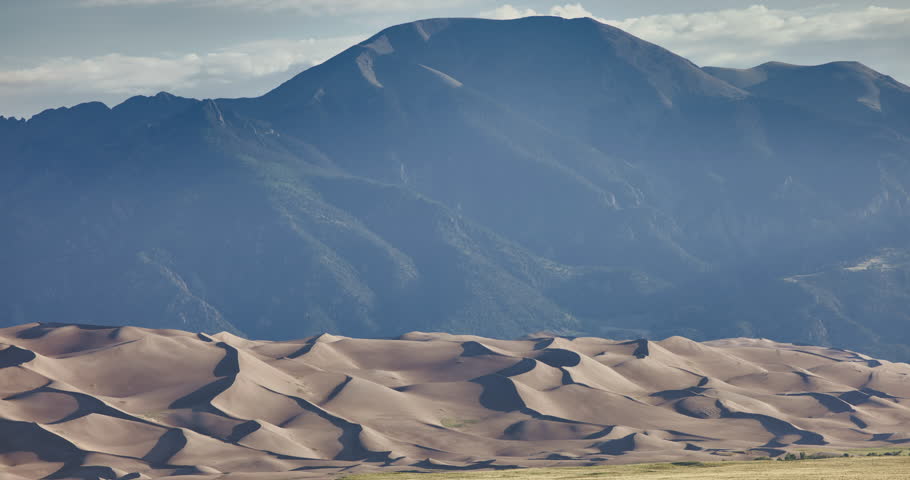 Telephoto time lapse panning shot of the Great Sand Dunes National Park and Preserve in Colorado during a summer daytime scene. Wind-shaped sand dunes stretch across the landscape with the Sangre de Cristo Mountains rising in the background as clouds move across the sky. Cinematic nature footage ideal for themes of travel, wilderness, conservation, climate, and the natural beauty of America’s national parks.