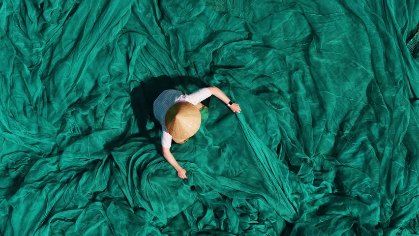 Aerial view Vietnamese fisherman with conical hat mending green fishing nets outdoors, traditional craft.