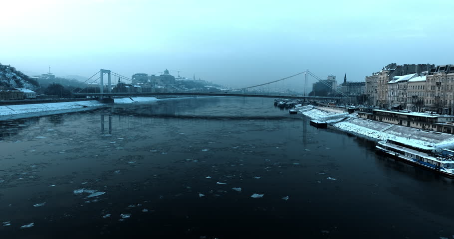 Elisabeth Bridge spanning the icy Danube River in Budapest during cold winter morning with snow-covered embankments and moody blue atmosphere.