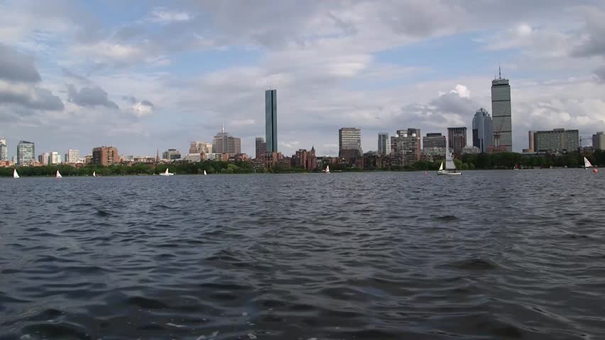 Boston skyline viewed across the Charles River on a partly cloudy day, with sailboats on the water and modern downtown skyscrapers in the distance.