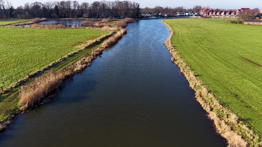 Aerial view of a calm canal running between grassy fields, with reeds along the bank, fenced pasture, scattered trees, and a small residential area in the distance.