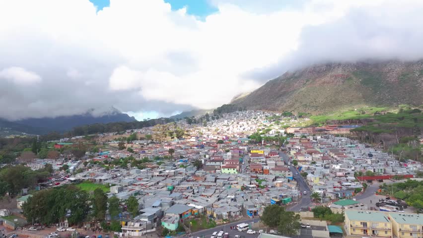 4K aerial drone footage of Imizamo Yethu township in Hout Bay, Cape Town, South Africa. Wide landscape view showing informal settlement against mountain backdrop with partly cloudy sky.