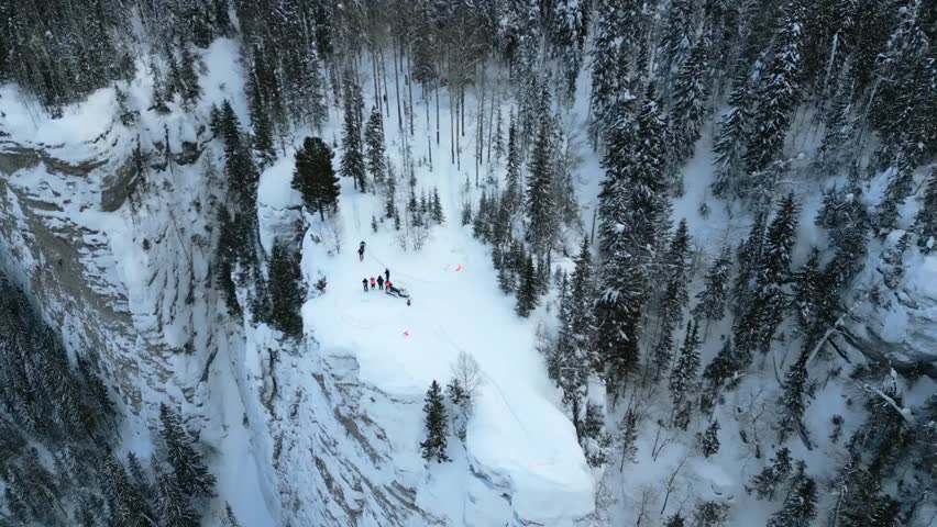 Aerial view of snow-covered mountain cliffs and fir trees, with tourists enjoying a winter day, showcasing frozen nature and adventure.